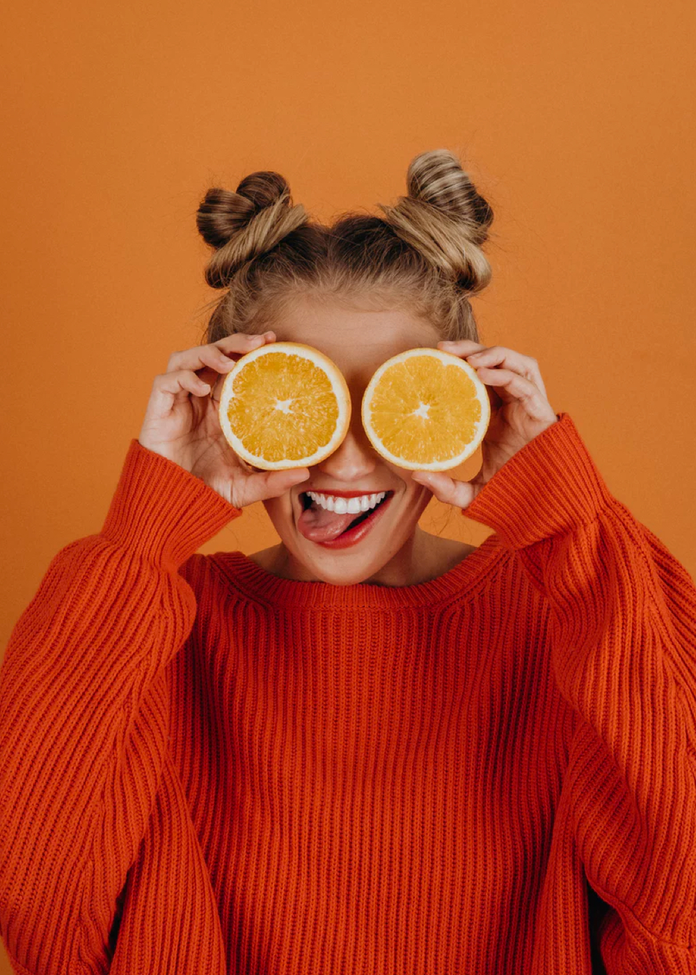 Sophie is standing in front of an orange background. She is smiling widely, sticking her tongue a little bit, and she's holding two halves of an orange in front of her eyes. She is wearing  cozy orange sweater and her hair is styled into top buns