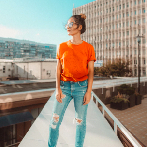 Sophie is posing ouside in an urban landscape. She is wearing a bright orange t-shirt, ripped blue jeans and sunglasses